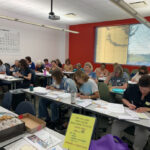 A photo of a classroom full of women writing on stacks of letters and envelopes.